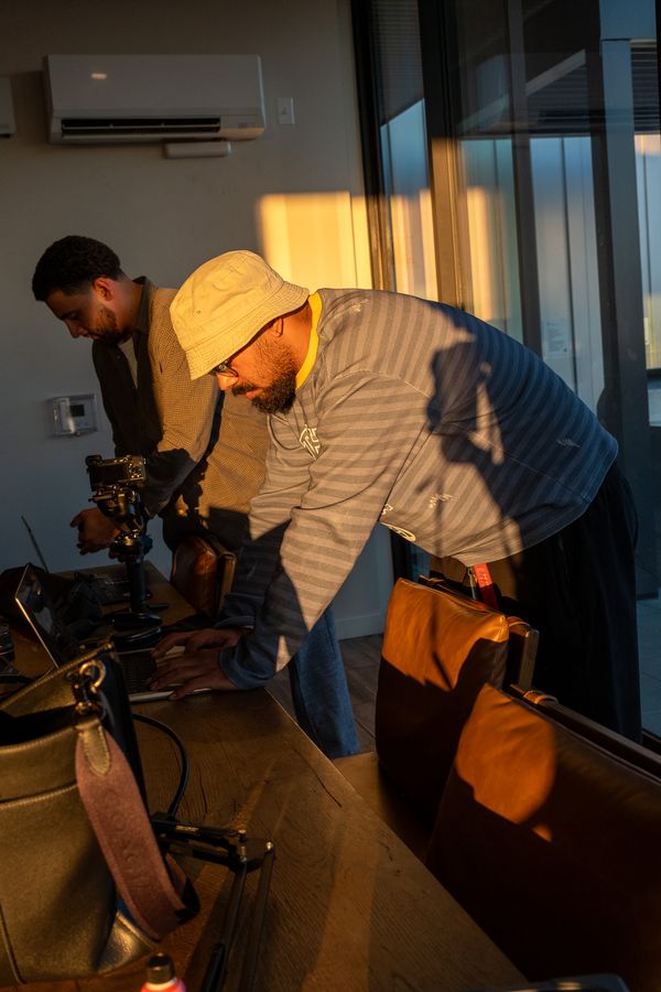 A man in a bucket hat leans over a laptop in a slice of golden sunset light while another checks a camera on a gimbal
