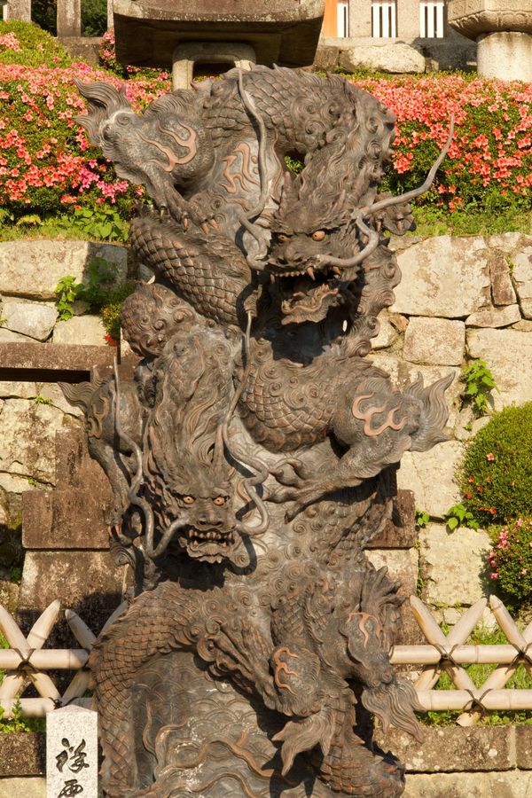 A bronze dragon statue coils in front of pink azaleas and a stone wall at a Kyoto temple