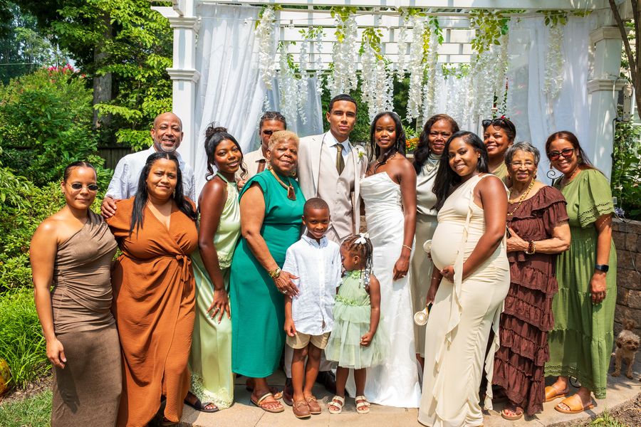 A large family in earth-tone dresses and suits gathered around the bride and groom under a white pergola