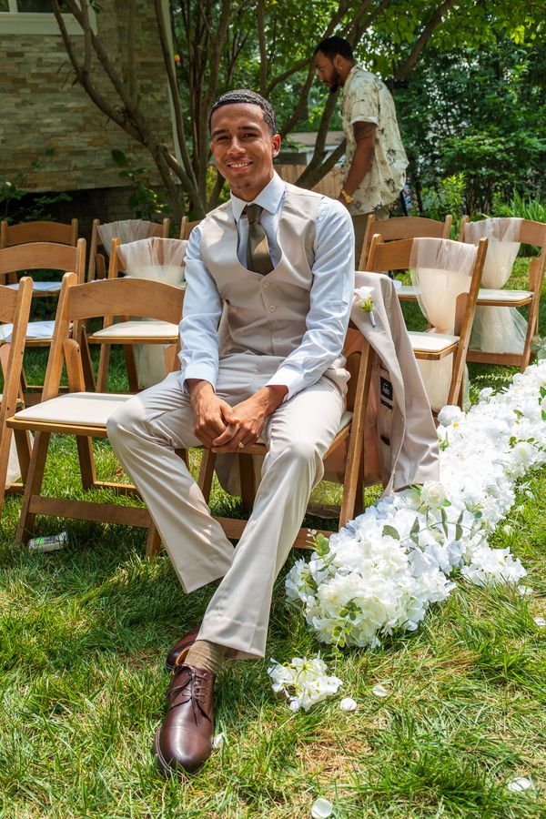 Groom in a cream vest and olive tie sits smiling on a wooden folding chair beside white flowers
